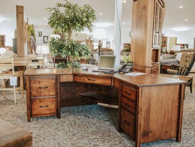 A wooden L-shaped executive desk with drawers, adorned with a laptop and phone, set in a cozy showroom featuring plush sofas and a large plant.